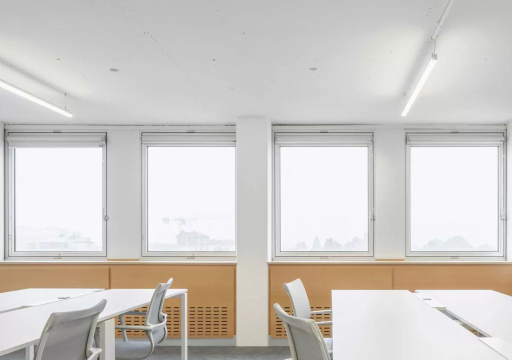 Salle de bureaux en open-space avec une grande luminosité, équipée de larges fenêtres et de mobilier blanc et minimaliste pour favoriser la concentration.